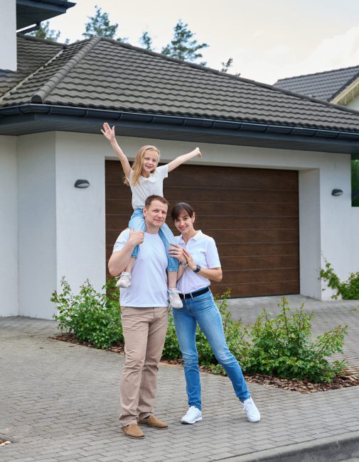 aaa A family of three posing in front of a house with a garage. The man carries the child on his shoulders, and the woman stands beside them, smiling. The child raises their arms enthusiastically.