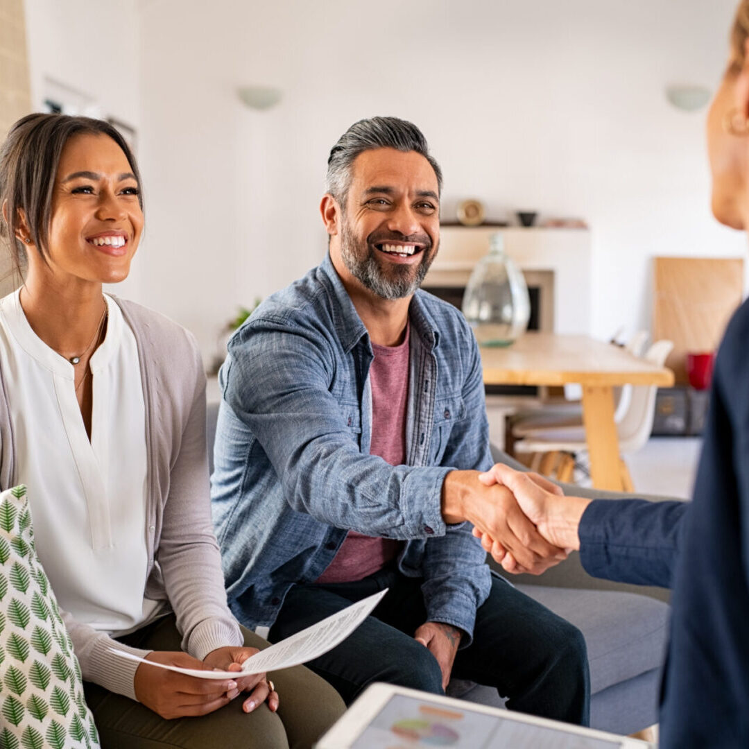 Multiethnic couple handshake with consultant at home A couple sits on a couch, smiling, as the man shakes hands with a person across from them in a living room setting.