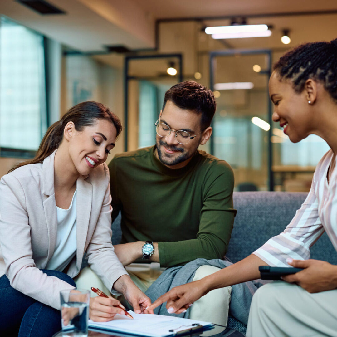 Happy couple signing an agreement with their insurance agent in the office. Three people sitting on a couch in an office setting, reviewing and discussing a document together.